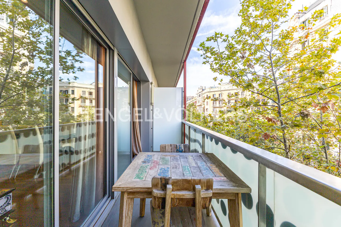Balcony with a wooden table and chairs overlooking a tree-lined street. Sliding glass doors lead inside.