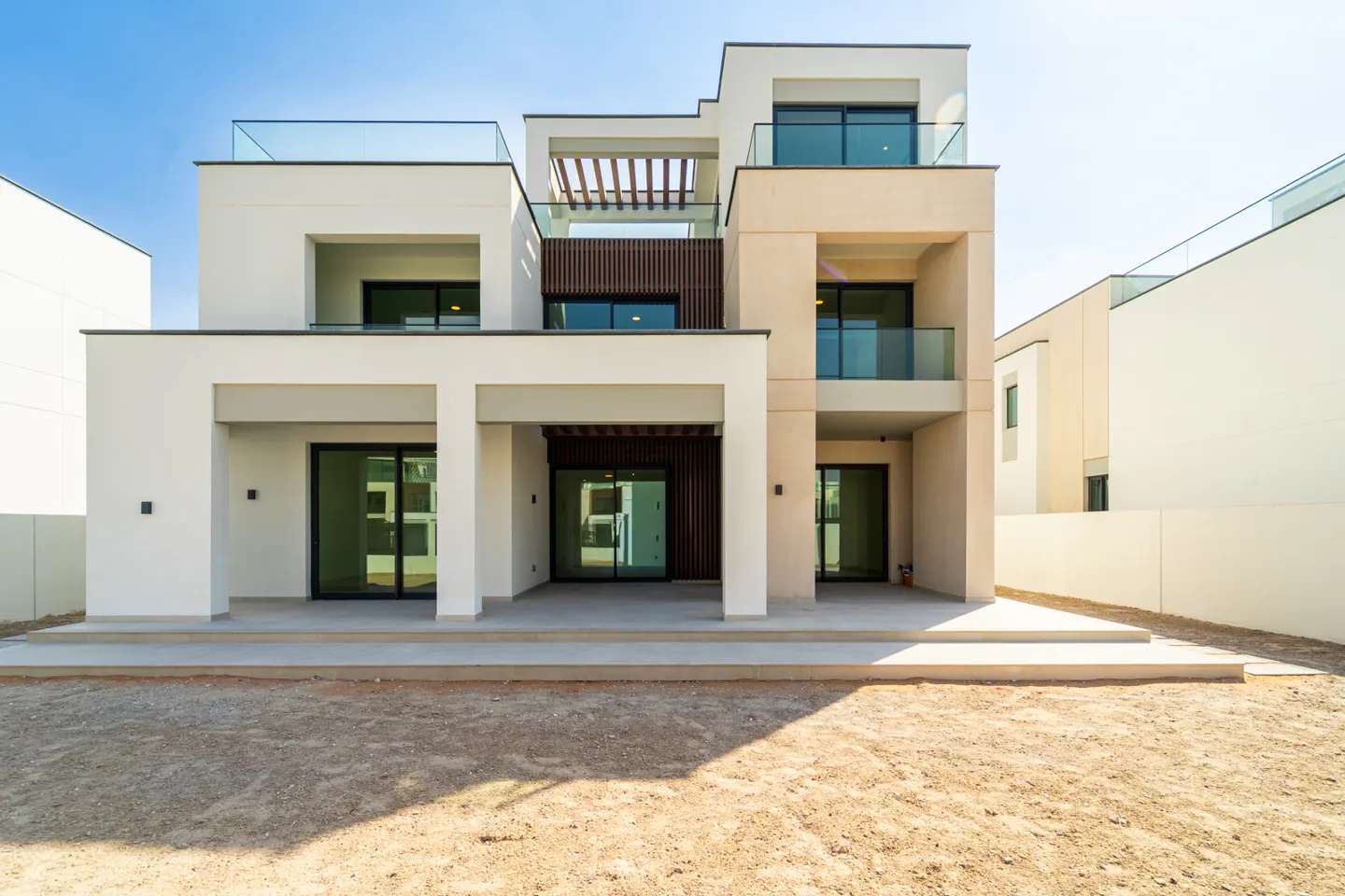 Modern two-story house with white and beige exterior, glass balconies, and a patio. The yard is dirt. Blue sky above.