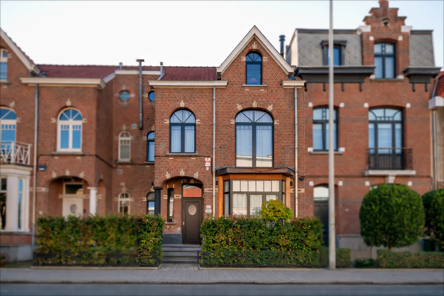 Red brick house with arched windows and a peaked roof, framed by green hedges and neighboring buildings.