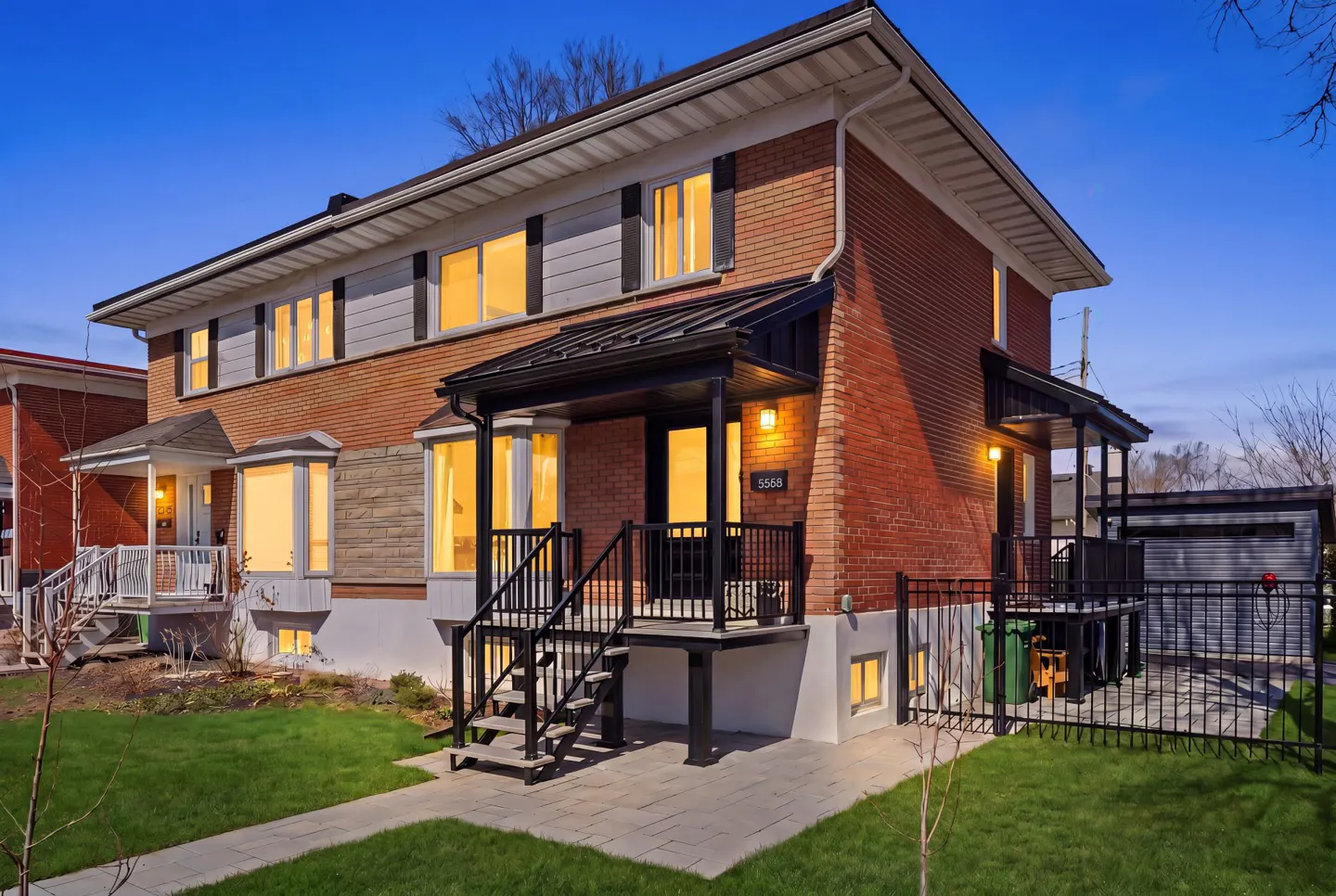 Exterior of a two-story red brick house with black railings and a small front yard at dusk.