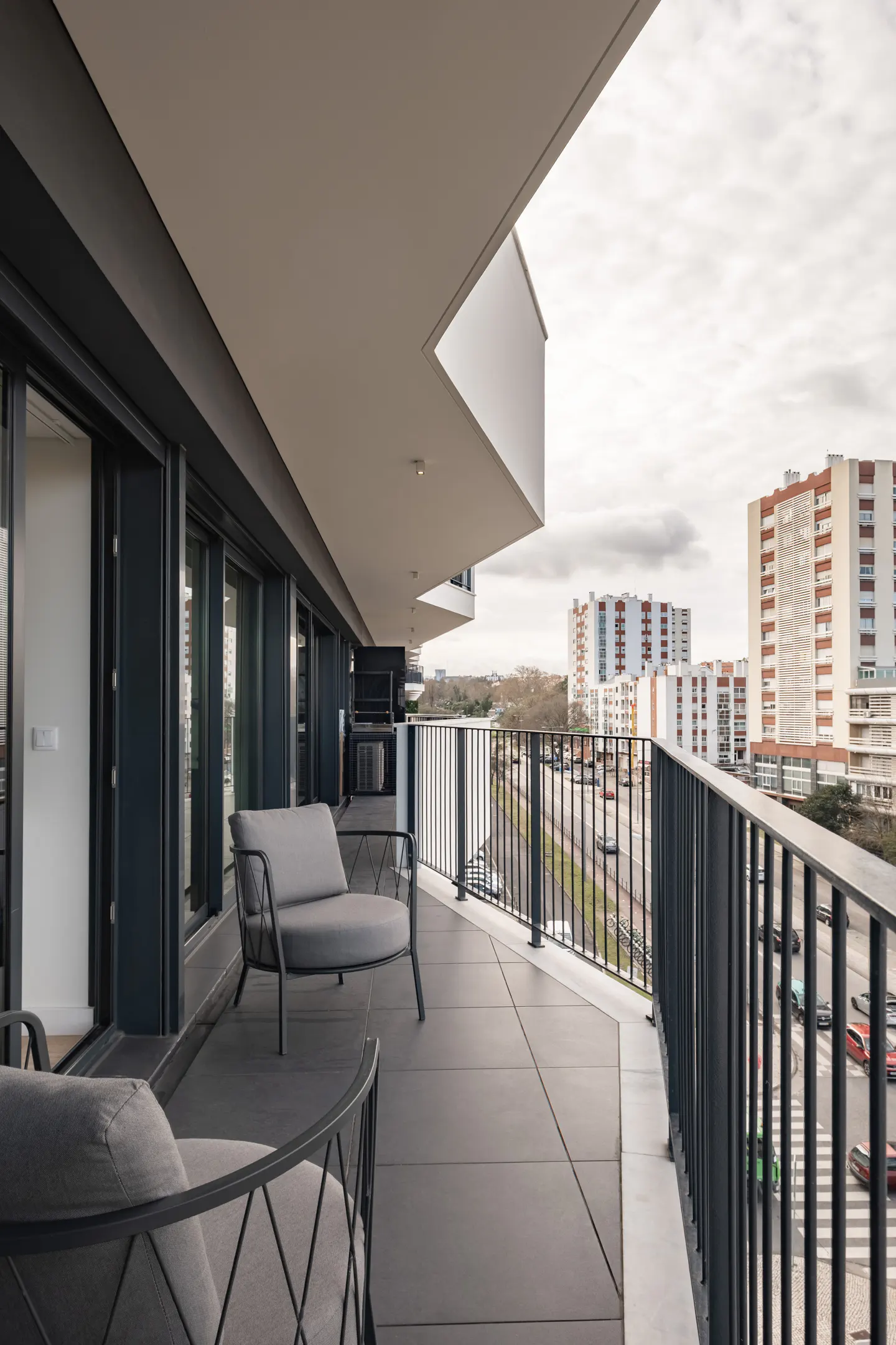 Modern balcony with gray lounge chairs, black railings, and city view. Cloudy sky above buildings and traffic.