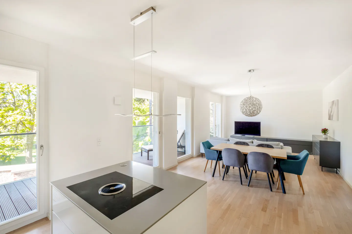 Bright, modern apartment interior with a kitchen island, dining table with gray and teal chairs, and a living room with a TV. Balcony visible.