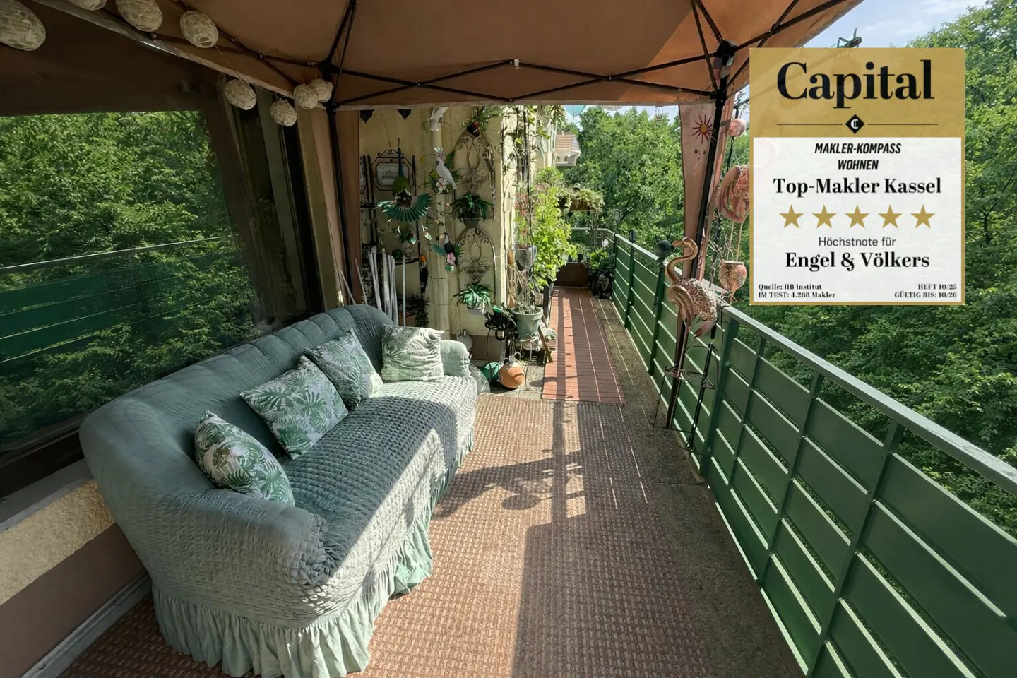 A cozy balcony with a green sofa, pillows, and a green railing overlooking lush trees. A "Capital" award for Engel & Völkers is visible.