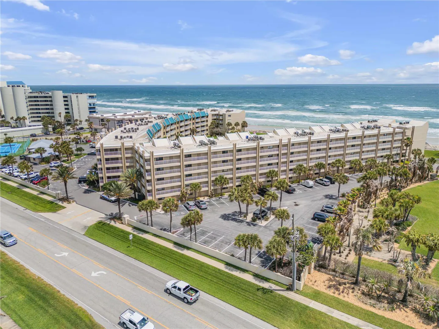 Aerial view of a beige condo building with a parking lot, palm trees, and the ocean in the background.