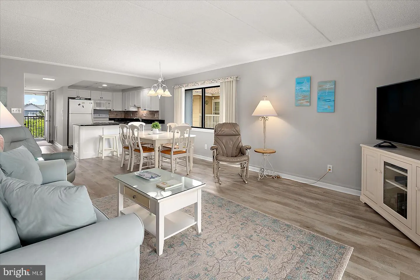 Bright, open-concept living space with a light blue sofa, white dining set, and a kitchen with white cabinets. A large window provides natural light.