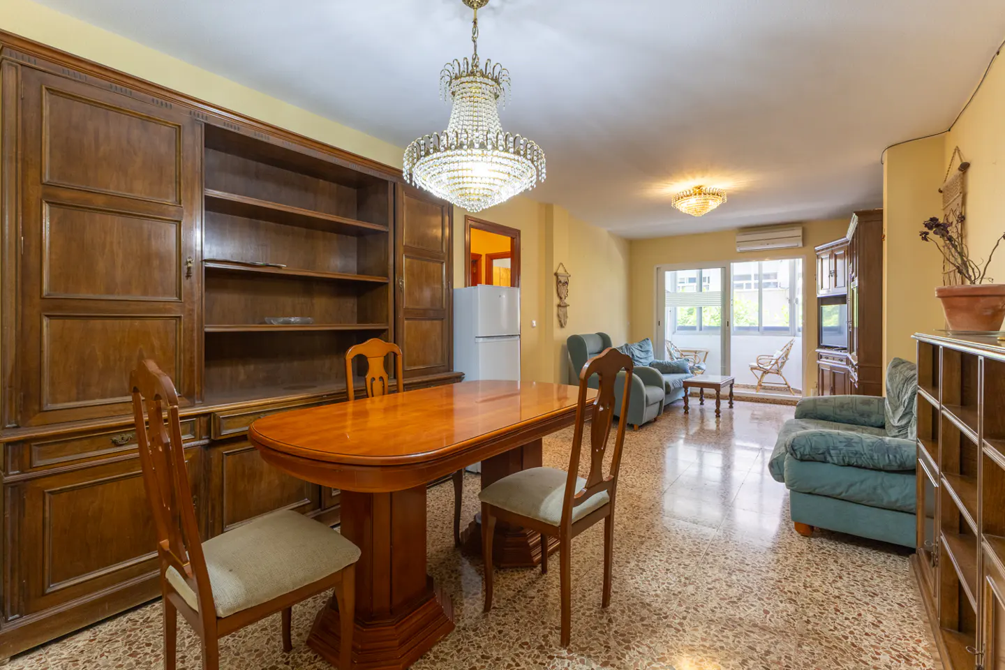 A living room with a wooden dining table, chairs, and a large wooden cabinet. A crystal chandelier hangs above the table. The room has a balcony and blue armchairs.