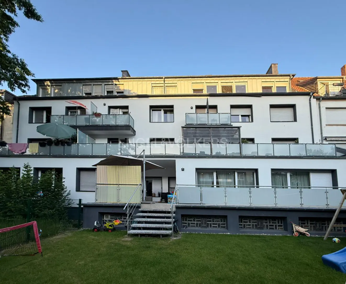 Exterior view of a three-story white apartment building with balconies and a green lawn. A soccer goal and toys are in the yard.