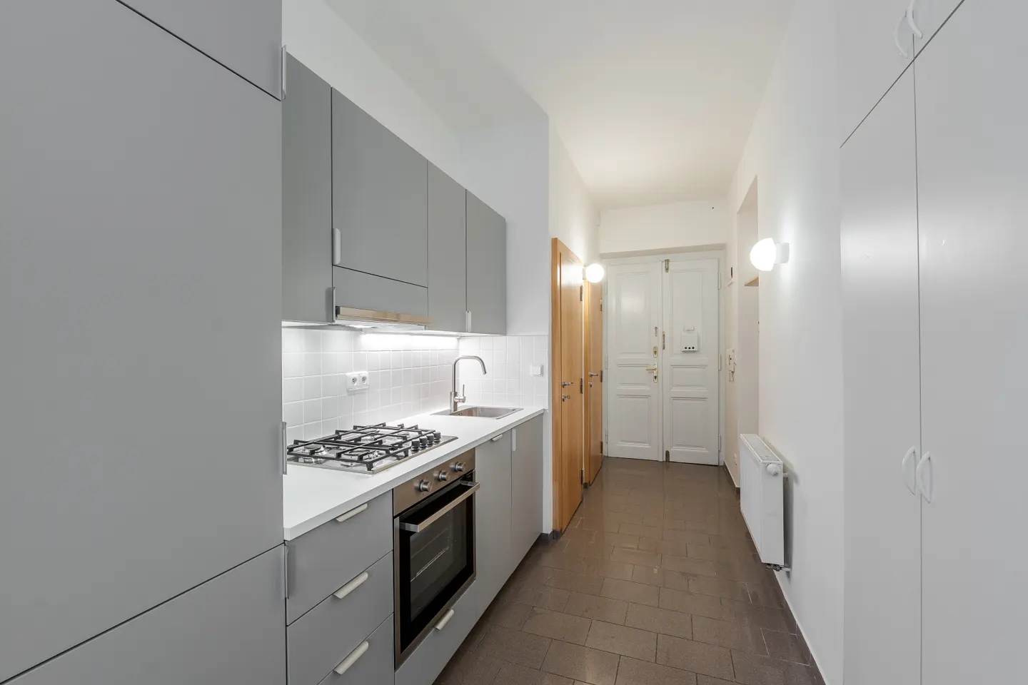 A modern kitchen with gray cabinets, white countertops, and stainless steel appliances. A hallway with white walls and brown tile floors leads to a white door.