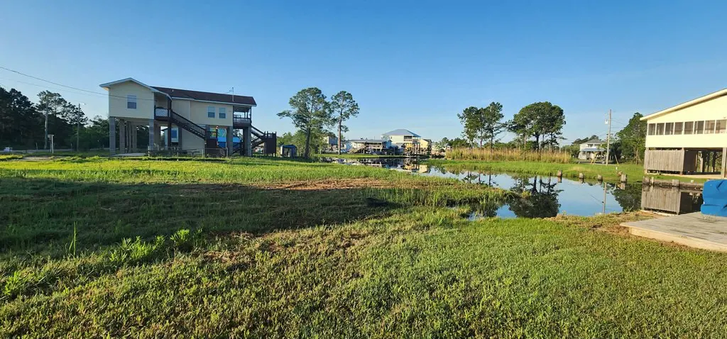 Waterfront property with stilt houses, green grass, and a canal reflecting the blue sky.