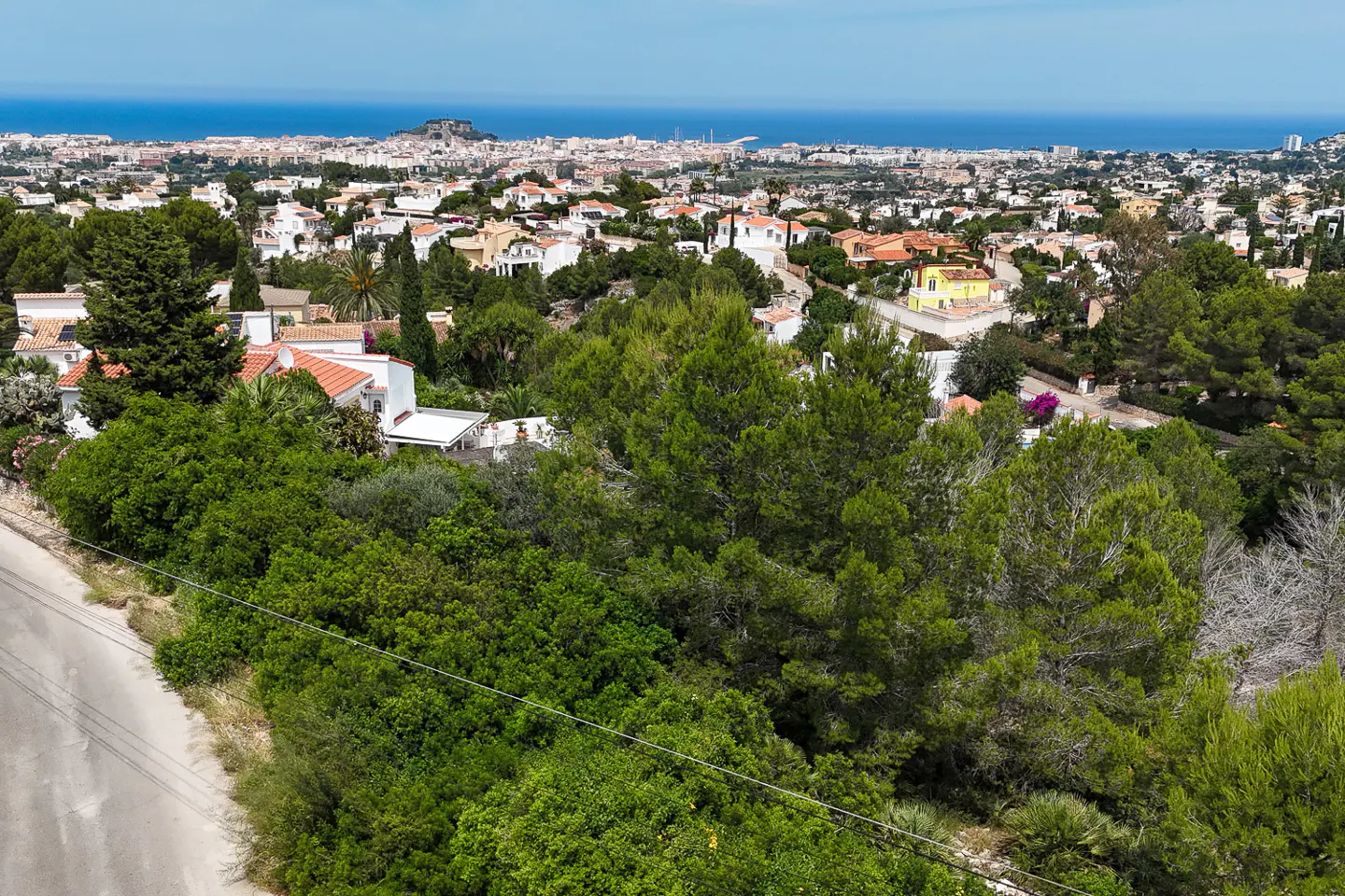 Aerial view of a lush green hillside with white villas overlooking a coastal town and the blue Mediterranean Sea.