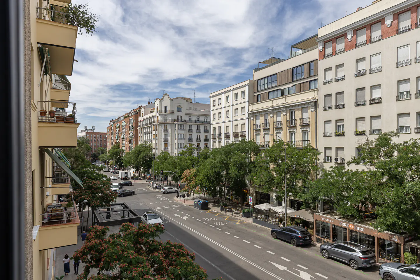 View from a balcony overlooking a sunny city street lined with historic buildings, green trees, and parked cars.
