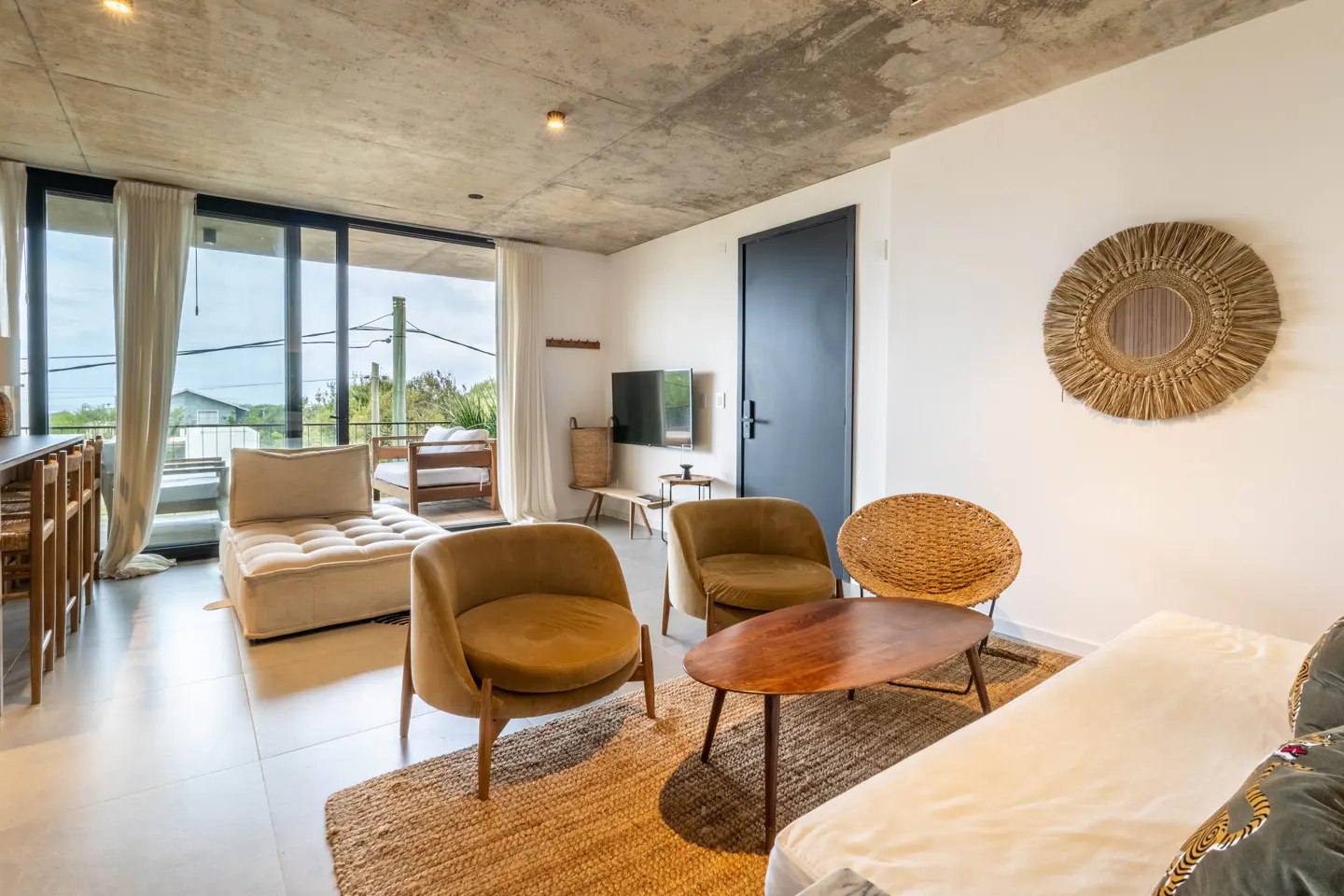 Modern living room with concrete ceiling, two velvet armchairs, a wooden coffee table, and a balcony with outdoor seating.