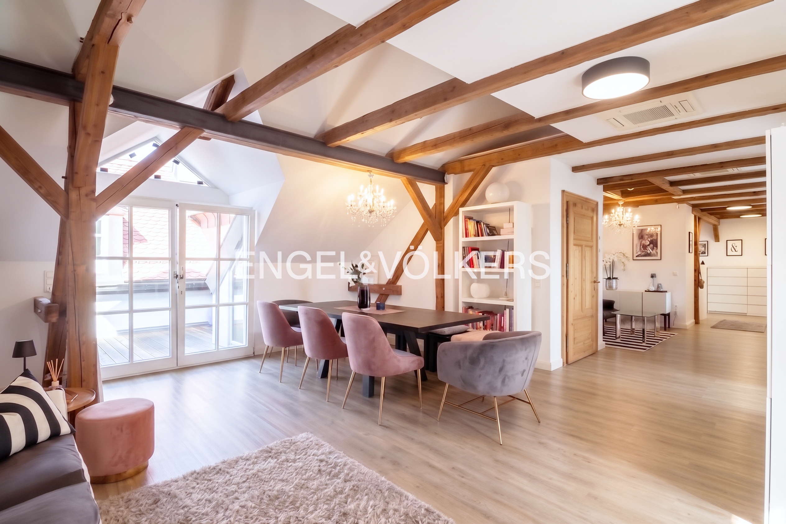 Dining area with wooden beams, pink and grey velvet chairs, a dark table, and a white bookshelf in a bright, open room. [AI-generated text]
