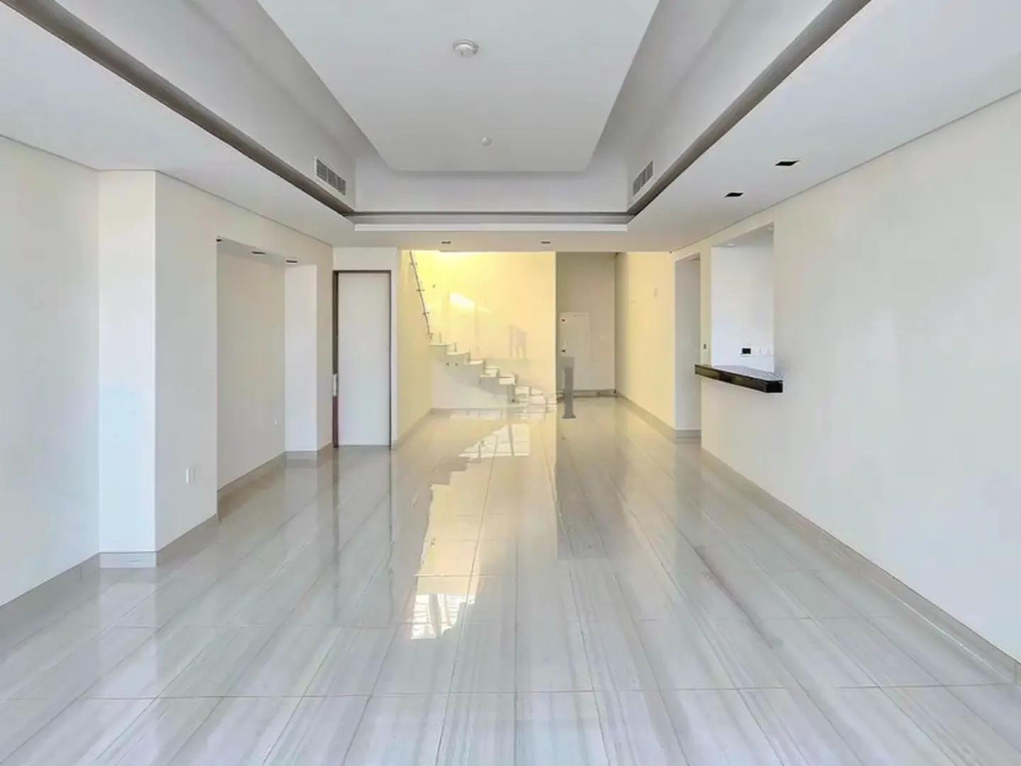 Bright, empty living room with white walls, tile floors, and a staircase with glass railings in the background.