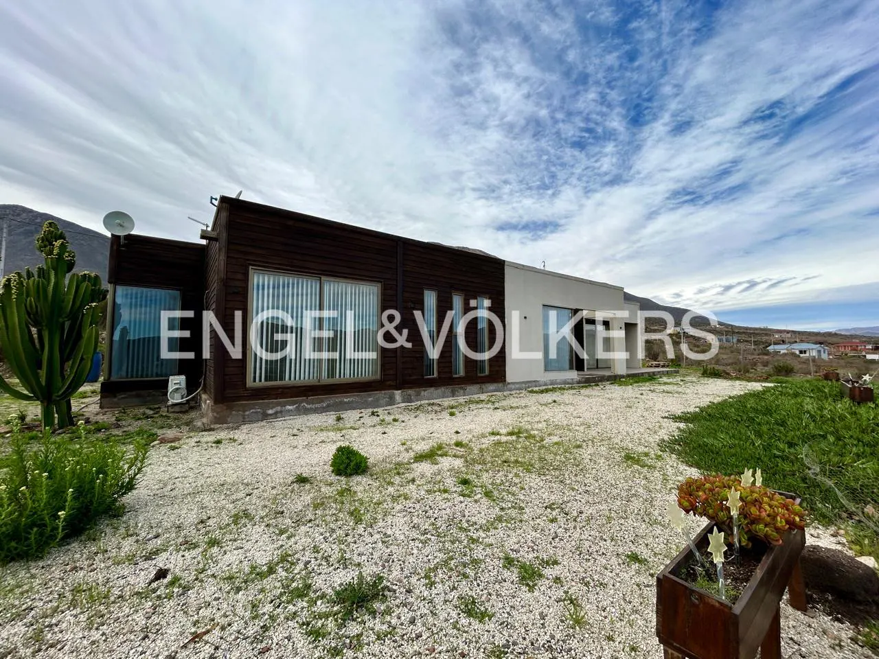 Modern home with dark wood and white exterior, gravel yard, large cactus, and mountain backdrop under a cloudy blue sky.