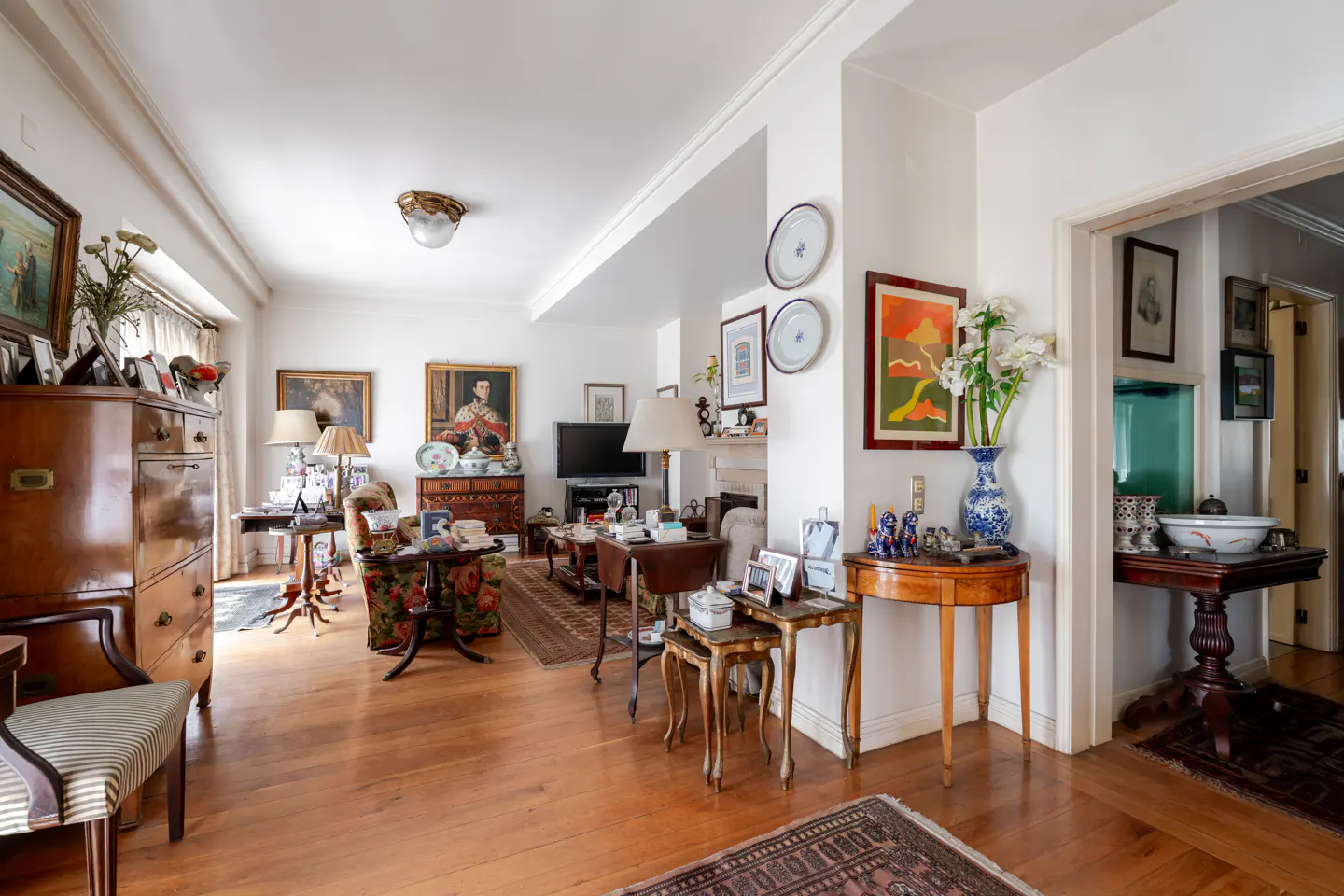 A living room with hardwood floors, white walls, and antique furniture. Paintings and decorative plates adorn the walls.