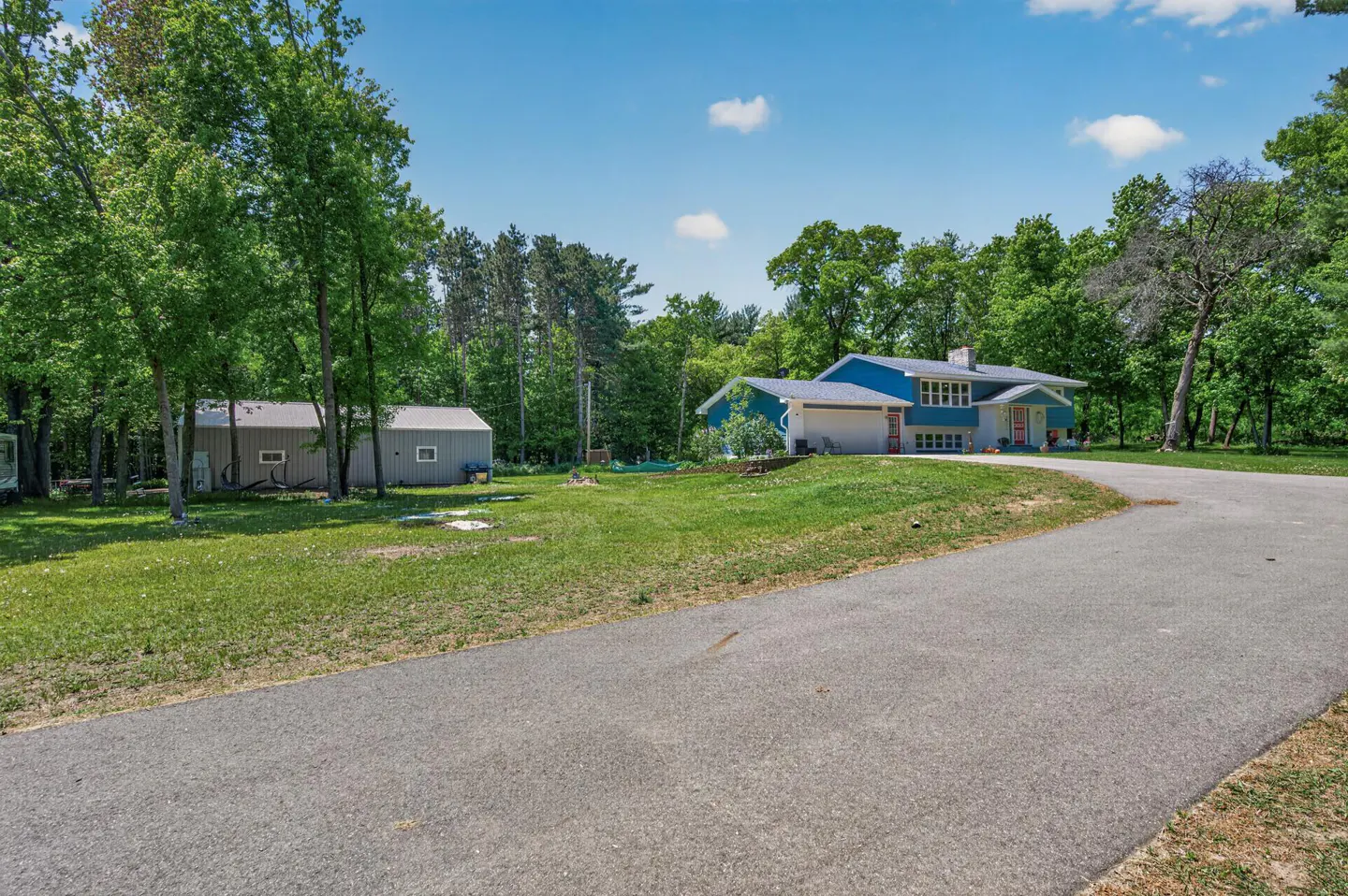 Exterior view of a blue and white house with a gray driveway and a gray shed surrounded by green trees.