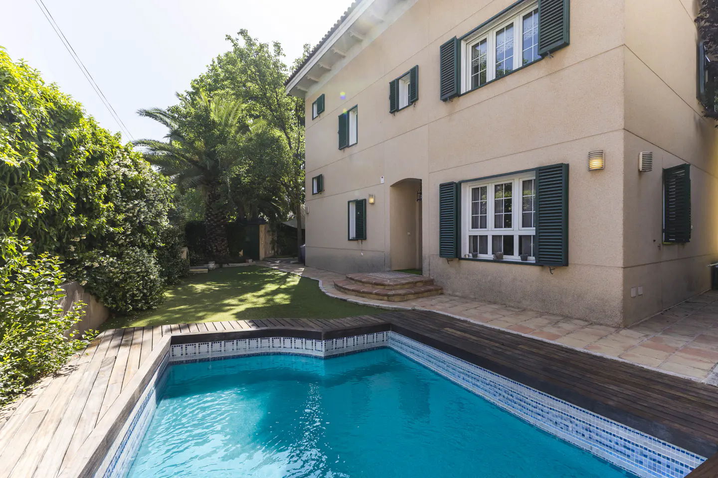 Two-story beige house with green shutters, a blue pool with a wooden deck, and green trees.