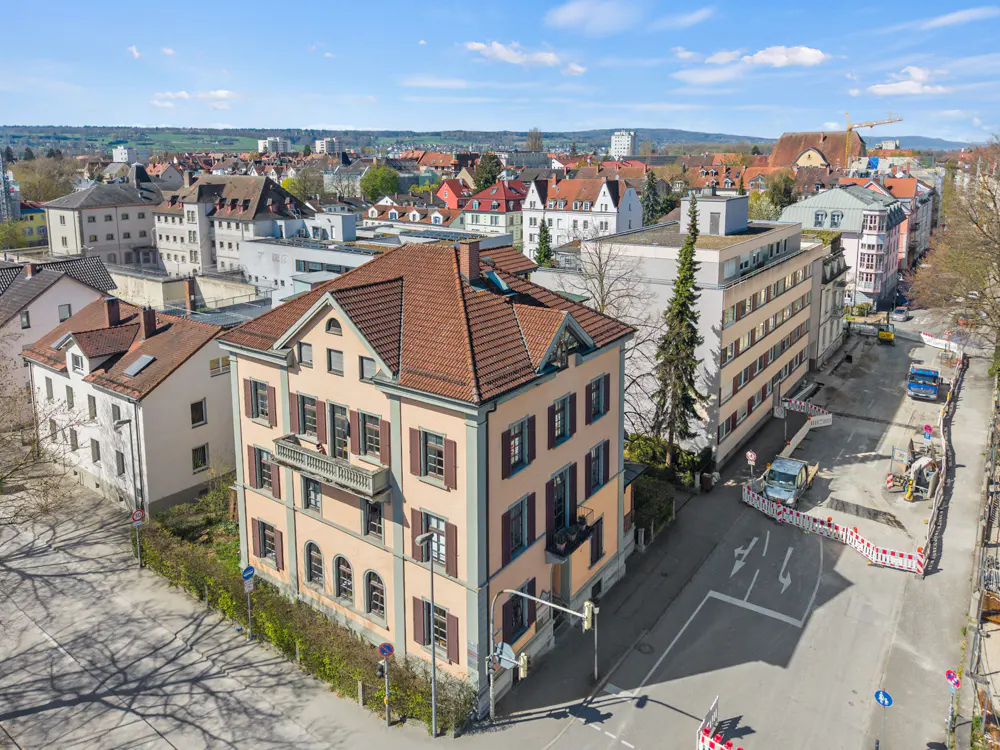 Aerial view of a peach-colored, three-story building with brown shutters and a red-tiled roof in a European city.