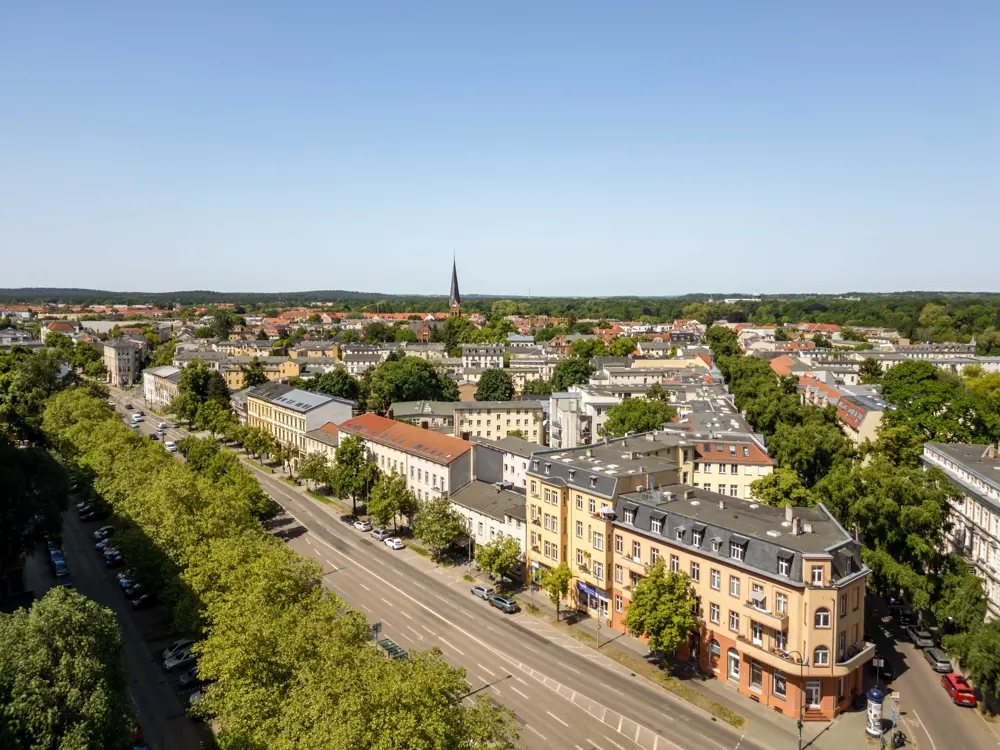Aerial view of a sunny European town with tree-lined streets, multi-story buildings, and a church spire in the distance.