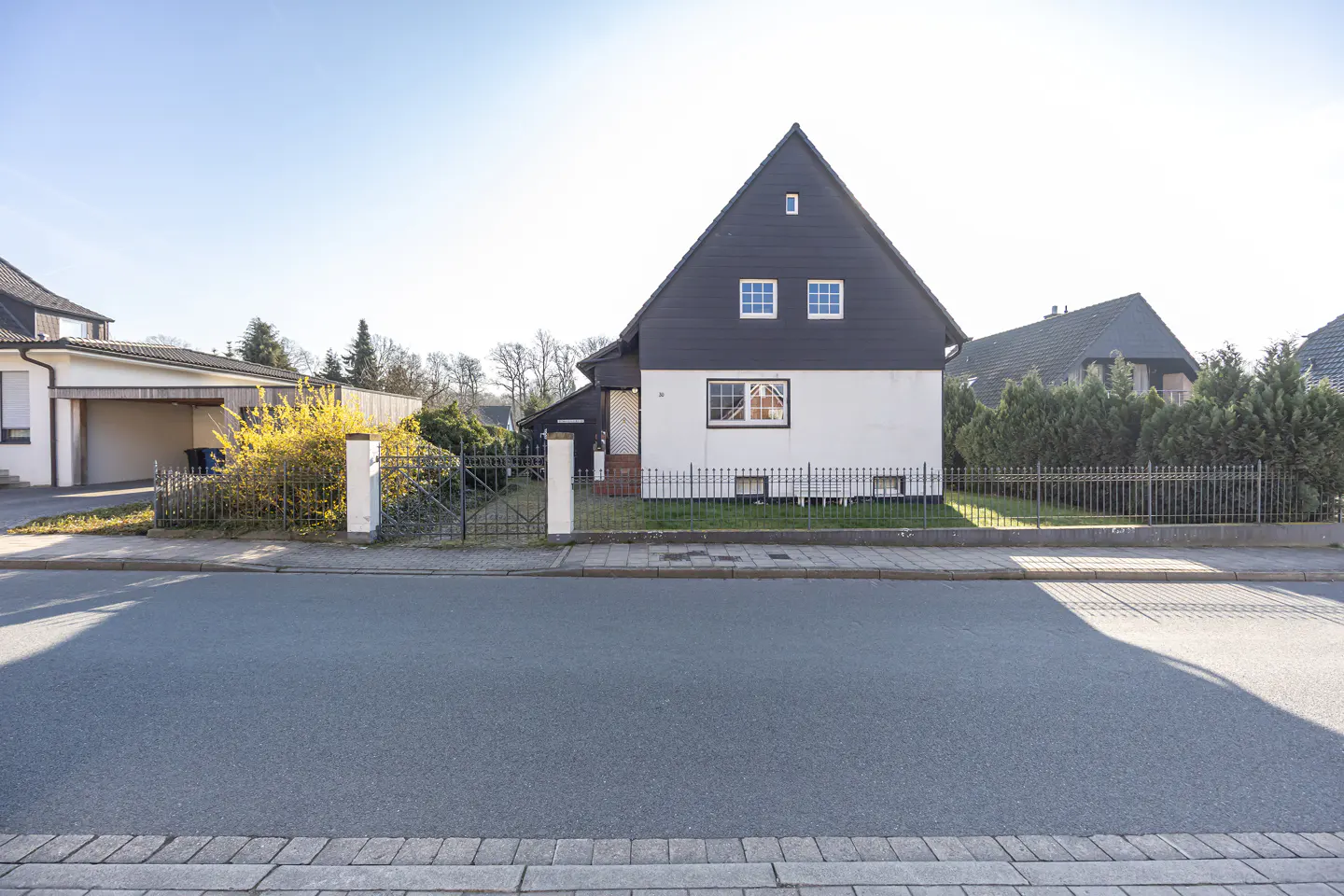 Two-story house with black roof and white siding, viewed from the street. A black metal fence surrounds the yard.