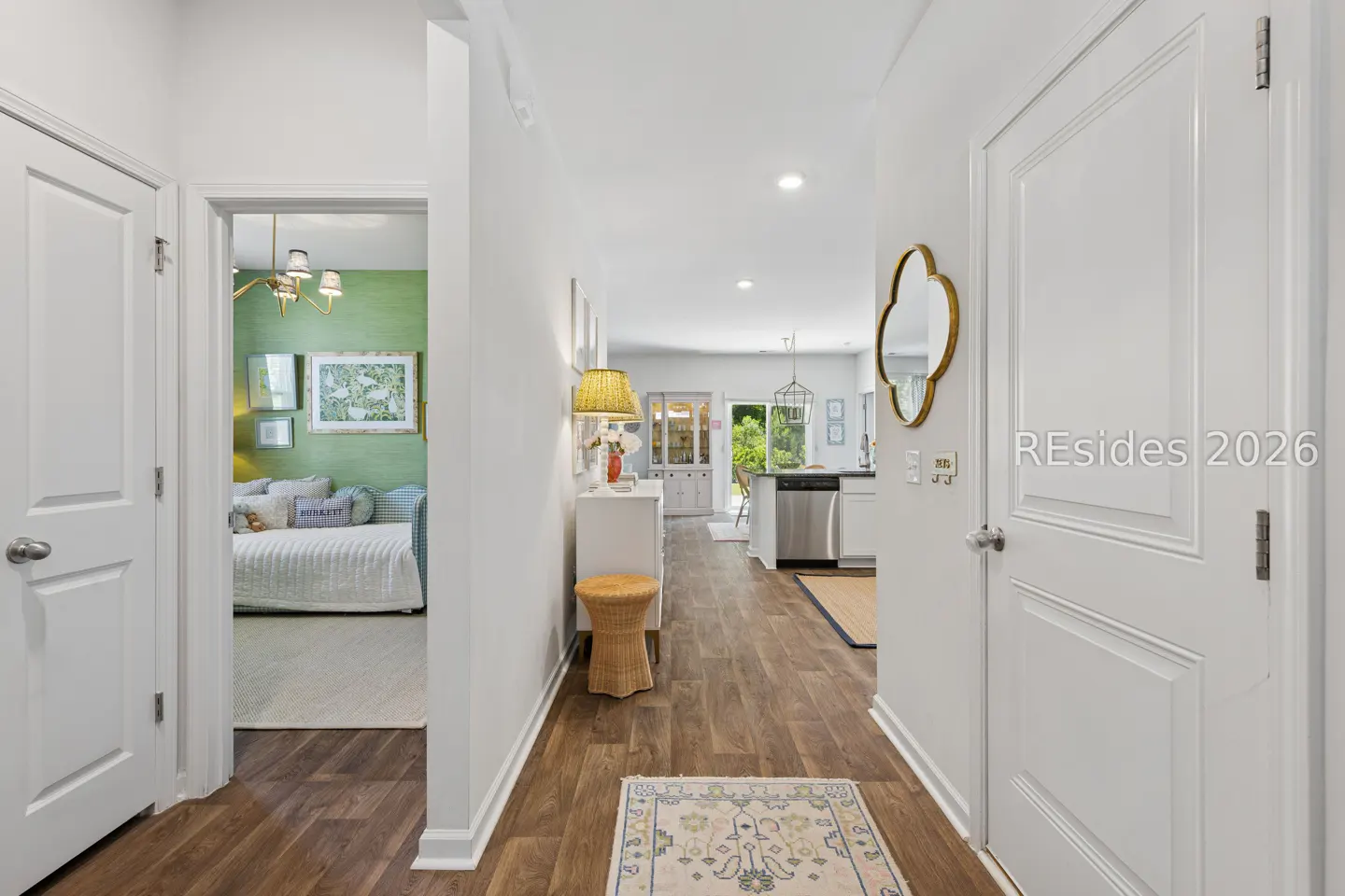 Hallway view with wood floors, white walls, and a glimpse into a green bedroom. A gold mirror hangs near a white door.