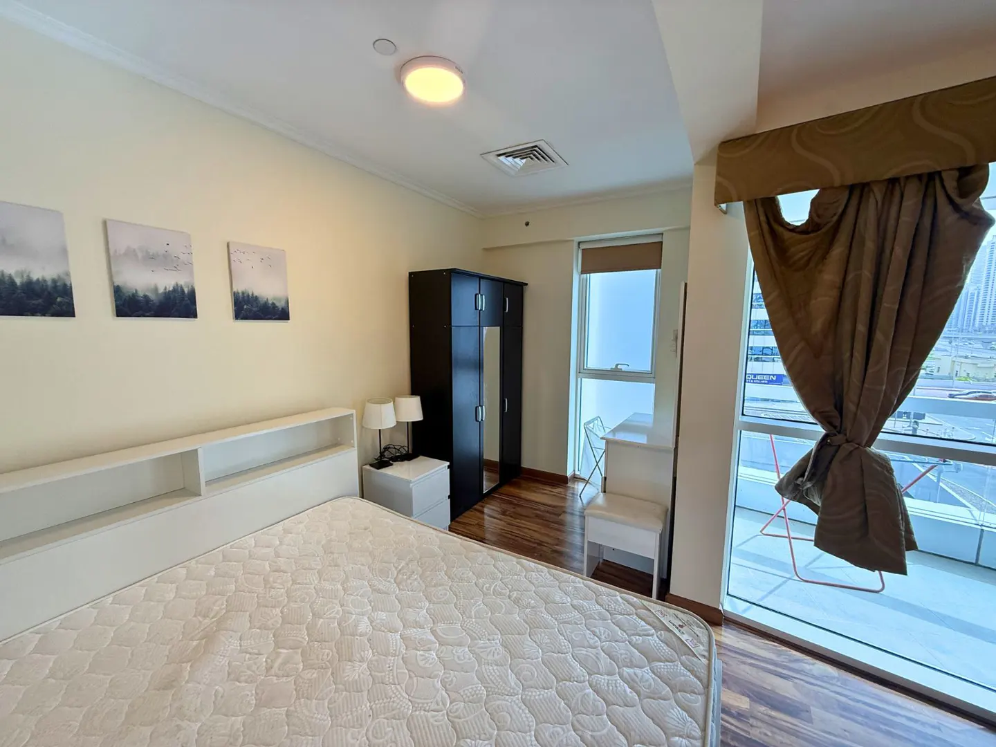 Bedroom with a bed, wardrobe, and desk. Three landscape paintings hang above the bed. A balcony is visible through a large window.