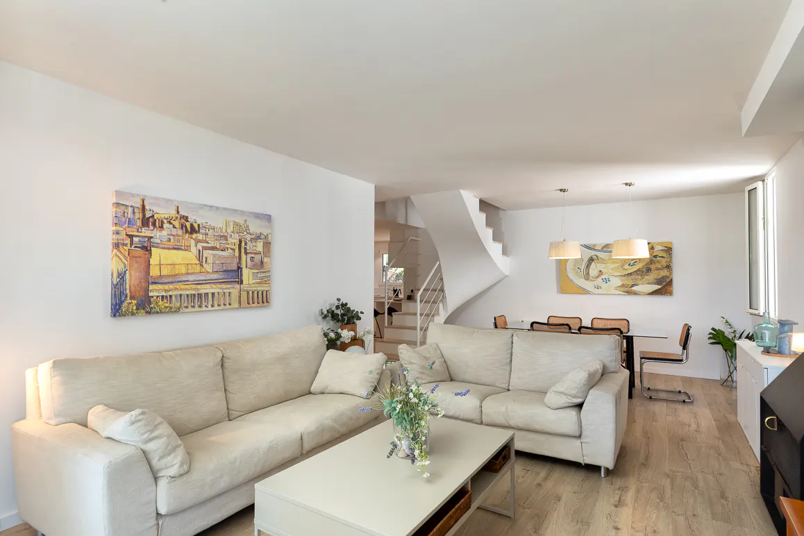 Bright living room with two beige sofas, a coffee table, and a dining area in the background with a curved staircase.