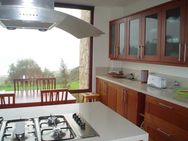 A modern kitchen with wood cabinets, a white countertop, and a stainless steel stove. A dining table sits near a large window with a view of greenery.