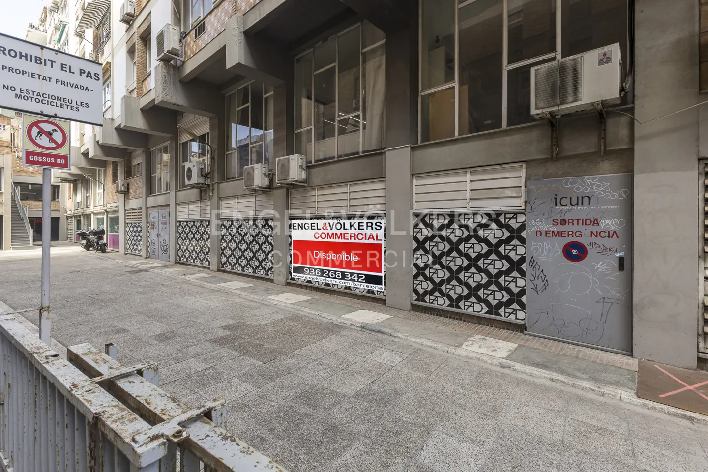 Exterior view of a commercial building with an Engel & Völkers "Commercial" sign. A "Prohibit El Pas" sign is visible. The building has air conditioning units and a gray emergency exit door.