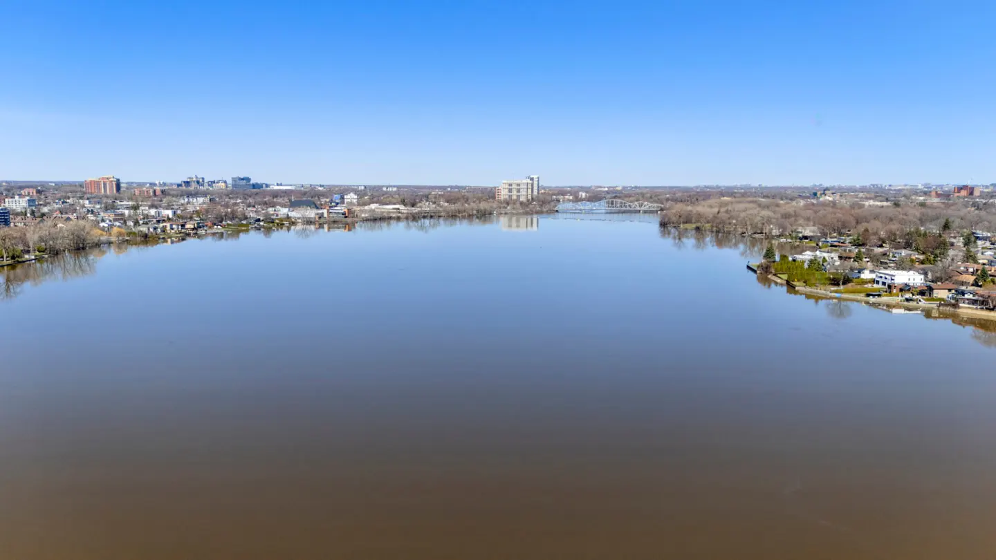 A wide river flows under a clear blue sky, reflecting the sky's color. Buildings and trees line the riverbanks.