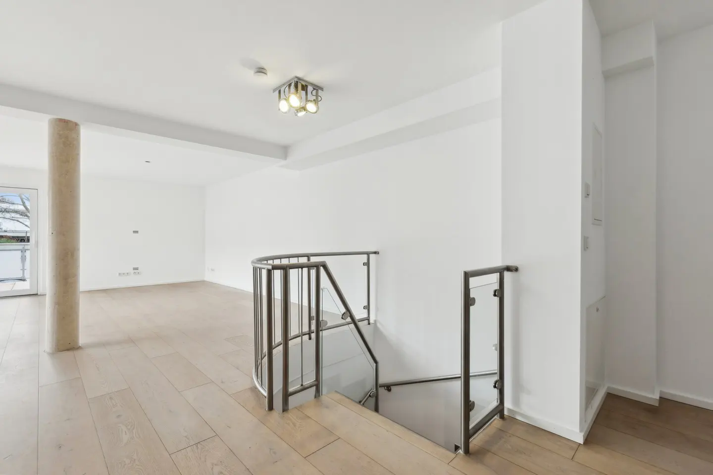 Bright, modern interior with light wood floors, white walls, and a curved staircase with glass railings. A beige pillar stands near a balcony door.