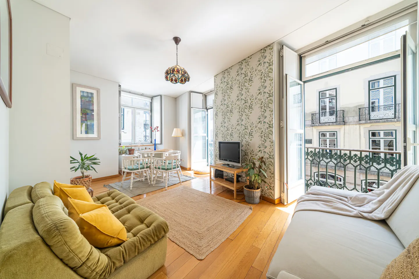 Bright living room with hardwood floors, olive sofa with yellow pillows, dining table, and balcony with building view.