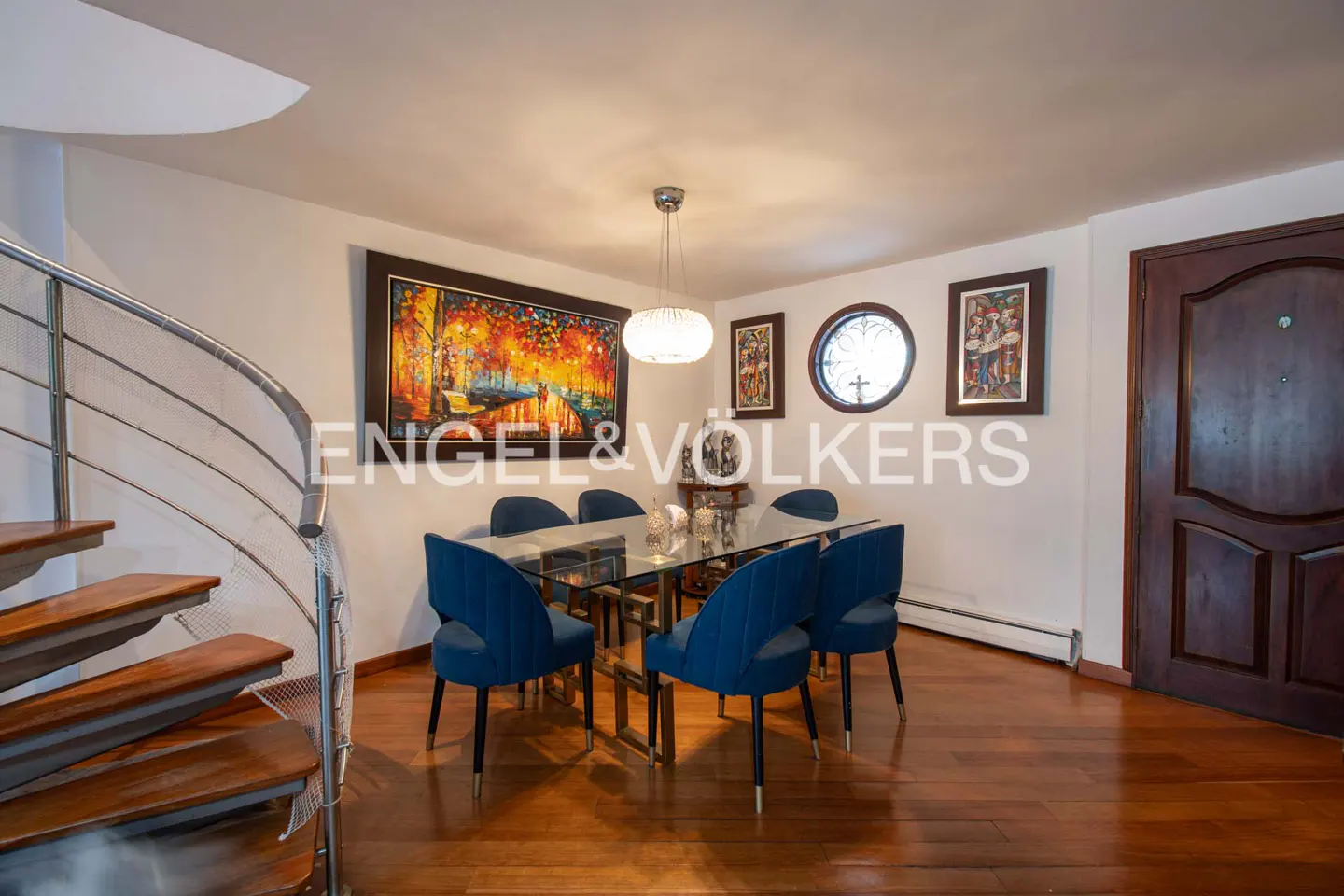 A dining room with a glass table, blue chairs, and a spiral staircase. Artwork adorns the walls, and a chandelier hangs above the table.