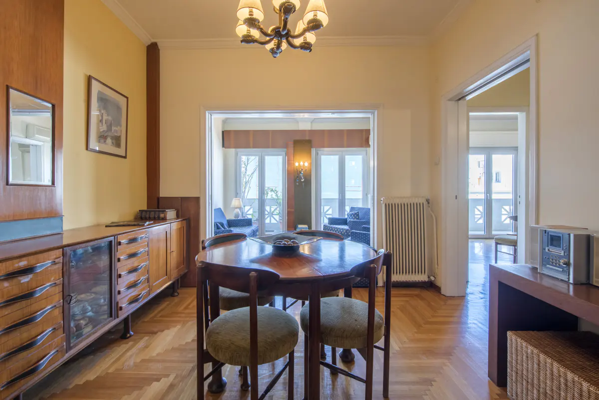 A dining room with a wooden table, four chairs, and a chandelier. A cabinet is on the left, and a doorway leads to a balcony with blue sofas.
