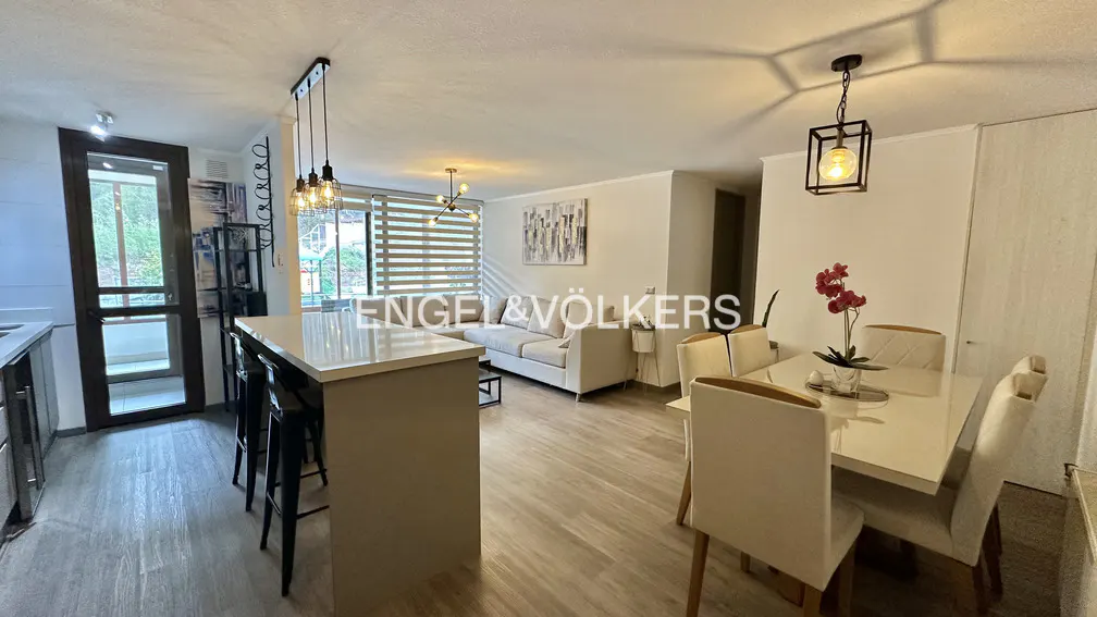 Open-concept living space with a kitchen island, dining table, and a cream-colored sofa. Natural light streams through the window blinds.