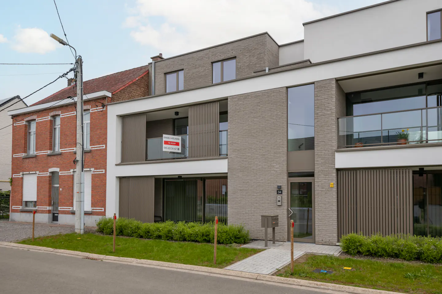Exterior view of a modern, multi-story apartment building with a "for sale" sign on the balcony. A brick house is visible on the left.
