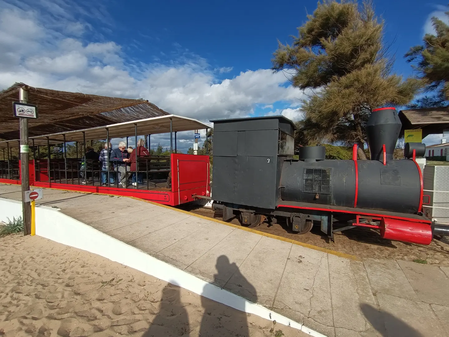 A black and red train with passengers is parked on a concrete platform next to a sandy area under a blue sky.