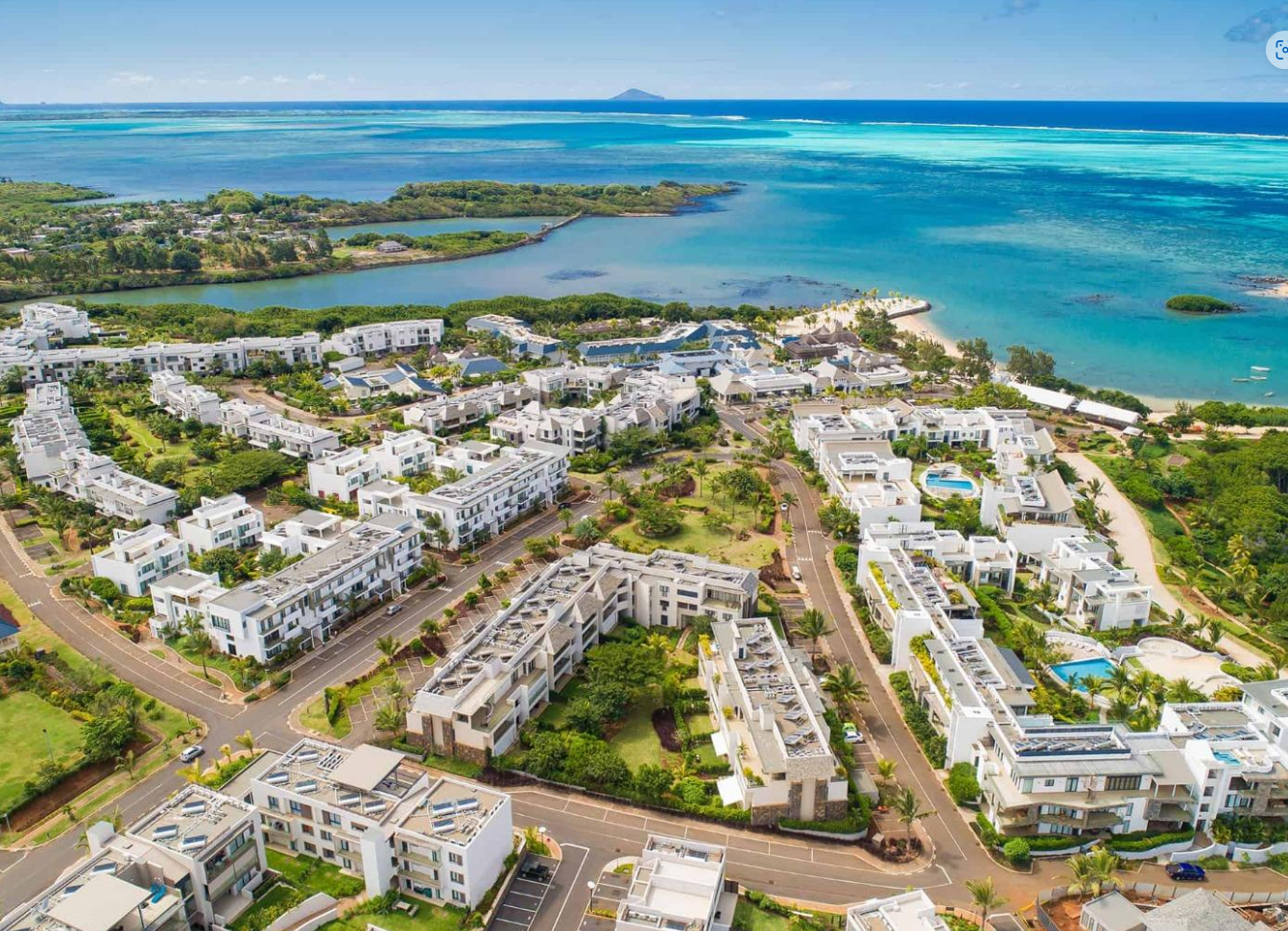 Aerial view of a coastal residential area with white buildings, green trees, and turquoise ocean under a blue sky.