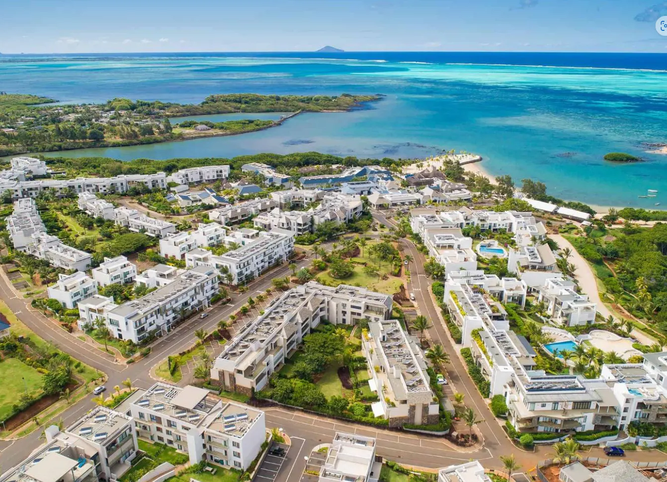 Aerial view of a coastal residential area with white buildings, green trees, and turquoise ocean under a blue sky.