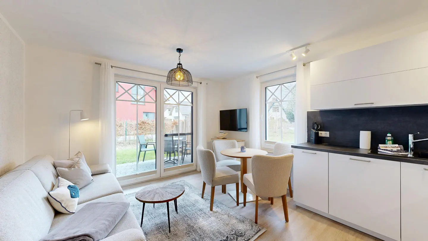 Bright living space with a sofa, round table with four chairs, and a modern kitchen with white cabinets. Patio doors lead to an outdoor seating area.