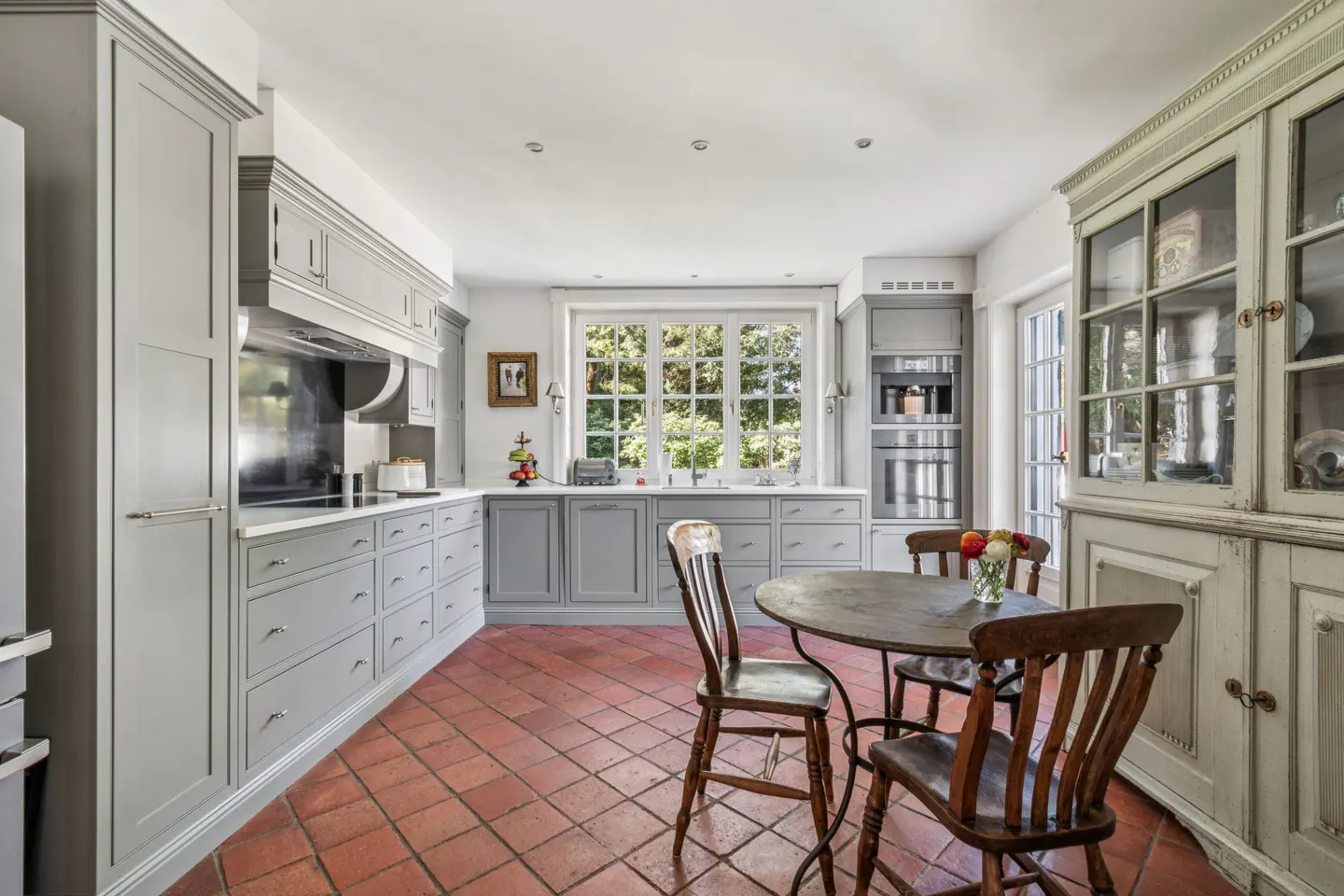 A kitchen with gray cabinets, terracotta tile floors, and a round wooden table with chairs. A large window overlooks greenery.