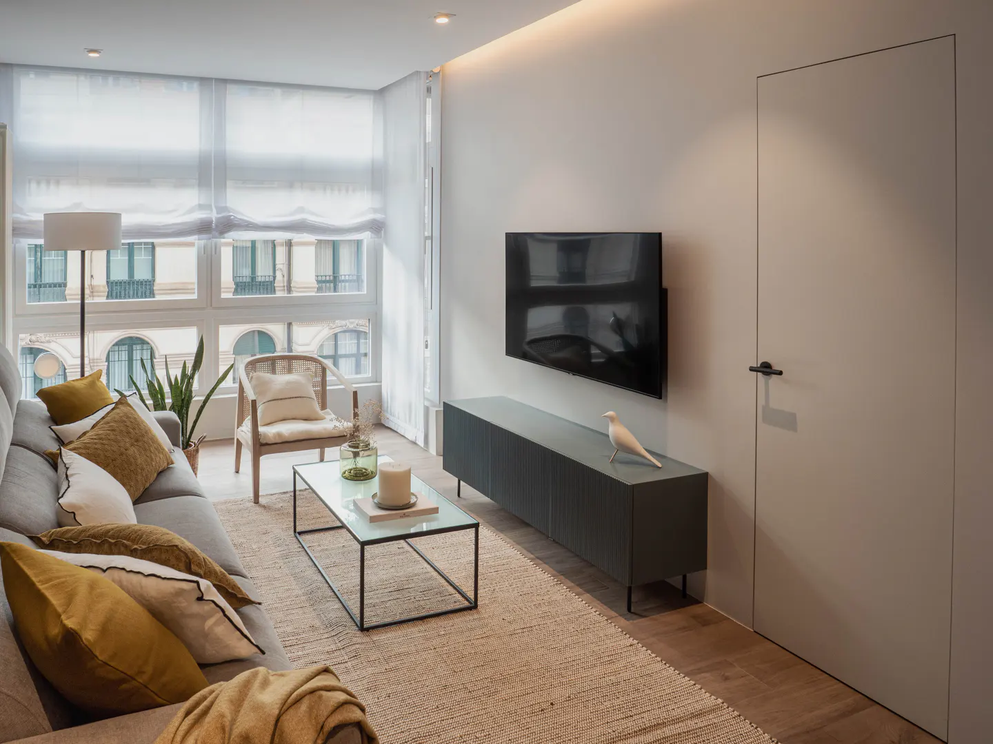 Living room with gray sofa, yellow pillows, glass coffee table, and TV. A white bird statue sits on a gray cabinet. Large windows with sheer curtains.