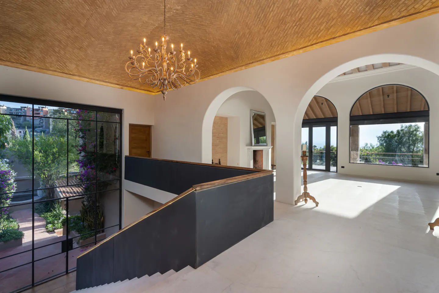 Interior view of a bright, modern home with white walls, arched doorways, and a brick ceiling. A chandelier hangs above a staircase with a black railing. Large windows offer views of lush greenery.