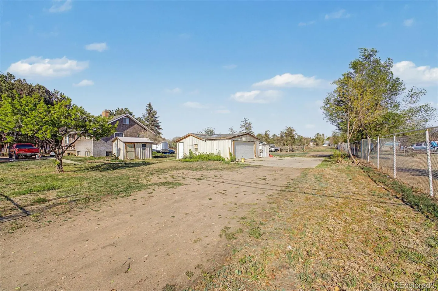 A rural property with a dirt driveway, a house, a shed, and a garage under a blue sky. A chain-link fence runs along the right side.