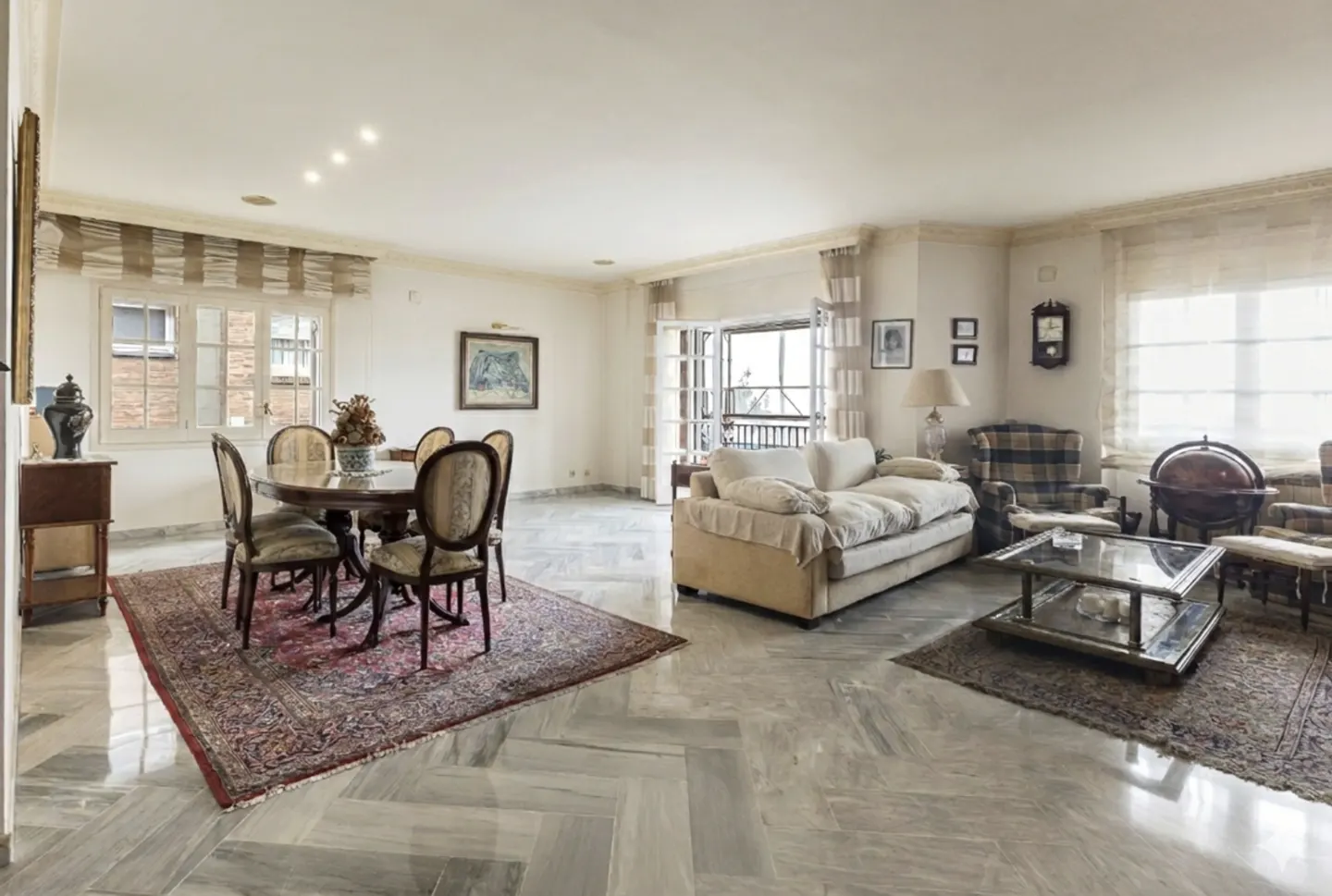 Spacious living and dining area with marble floors, a round table with chairs, and a beige sofa. Natural light streams through the windows.