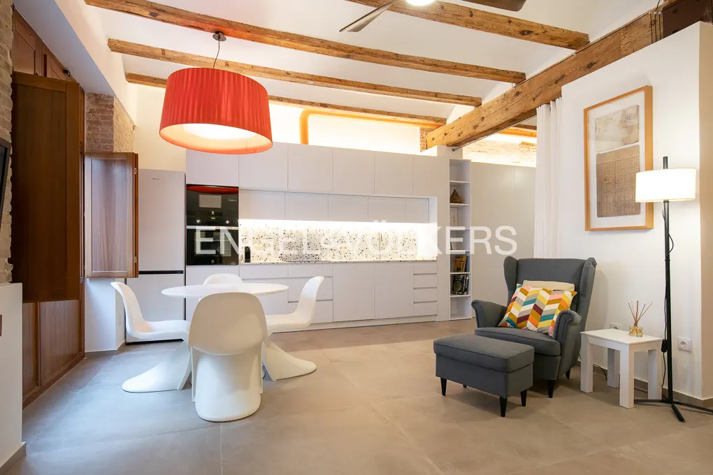 Open-concept living space with white kitchen, round table, and gray armchair with ottoman. Exposed wooden beams on the ceiling.