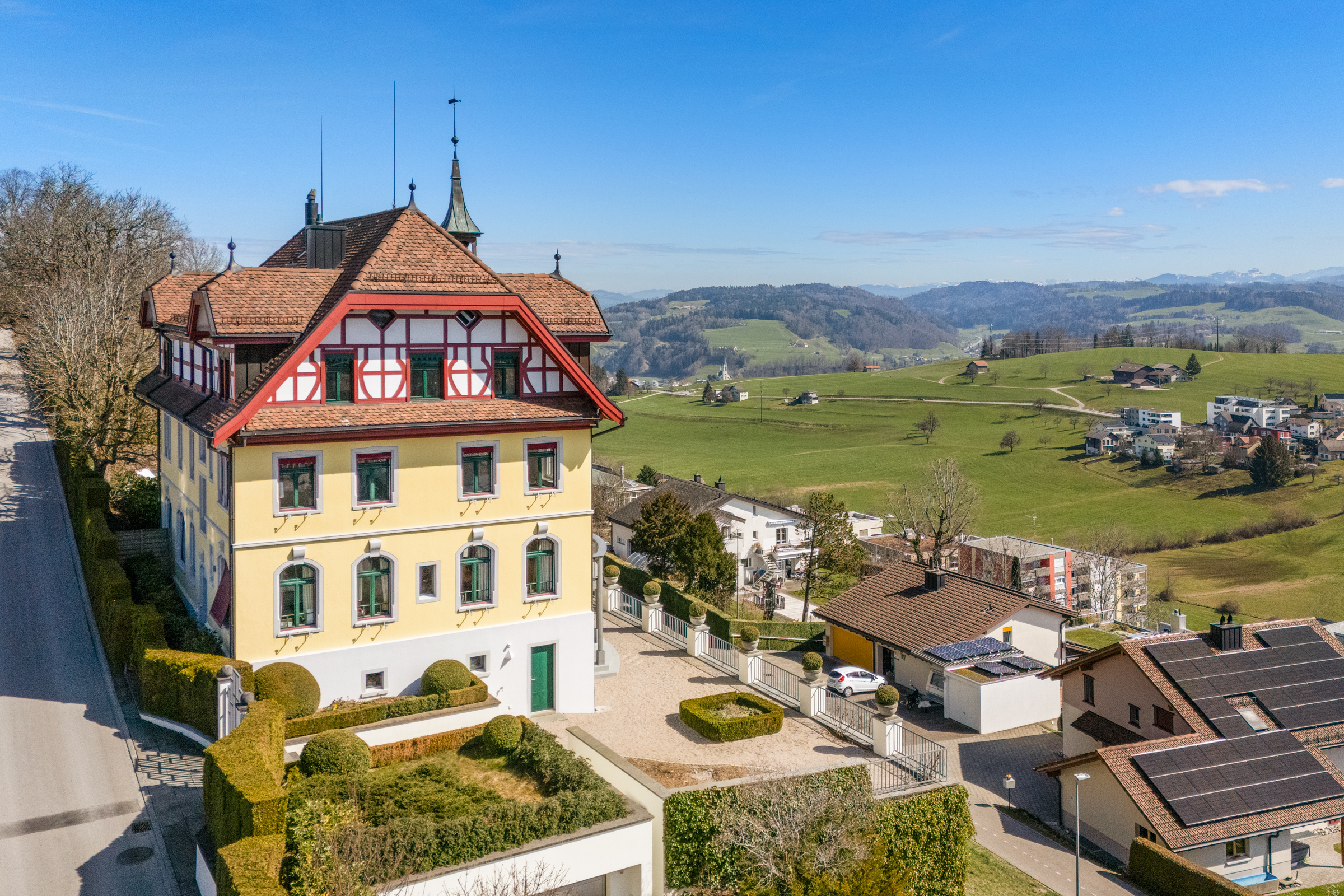 Aerial view of a yellow, three-story house with a red roof, green trim, and a decorative white and red half-timbered top floor. Rolling green hills in the background.