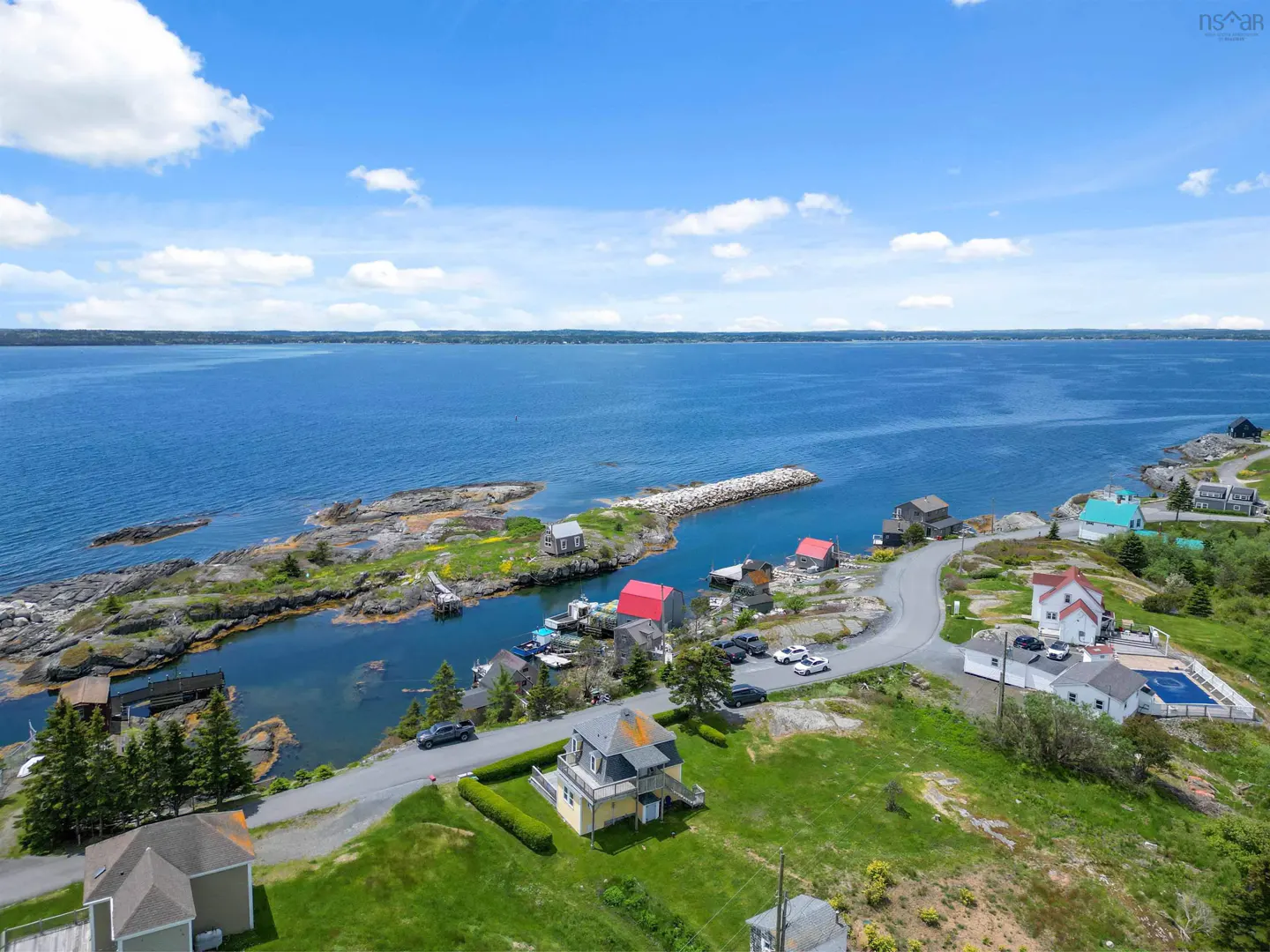 Aerial view of a coastal village with houses, a winding road, and a rocky inlet along a deep blue ocean under a sunny sky.