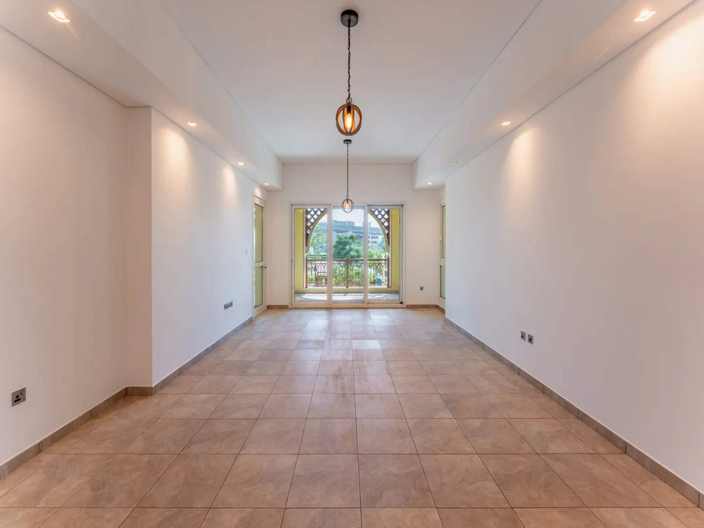 Bright, empty room with beige tile floor, white walls, and recessed lighting. Balcony with sliding glass doors at the far end. Two hanging pendant lights.